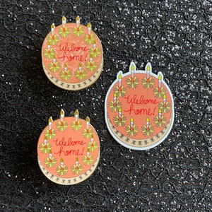May include: Three round enamel pins featuring a peach-colored cake design with yellow flowers and lit candles. The text Welcome home! is written in red cursive in the center of each cake. The pins have a gold-toned metal border and edge.