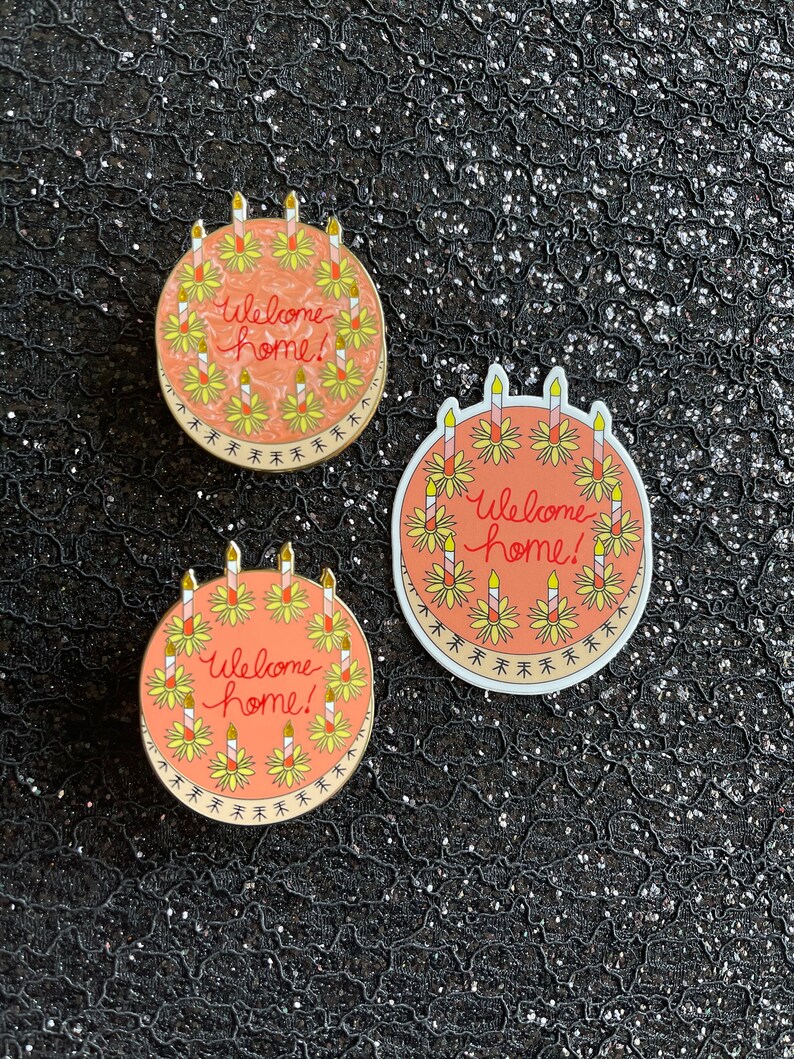 May include: Three round enamel pins featuring a peach-colored cake design with yellow flowers and lit candles. The text Welcome home! is written in red cursive in the center of each cake. The pins have a gold-toned metal border and edge.
