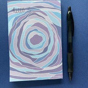 May include: A blue and purple patterned notepad with the text "hello :)" written on it. The notepad is on a blue surface with a black pen next to it. The notepad has the text "Fortuna Pins" at the bottom.