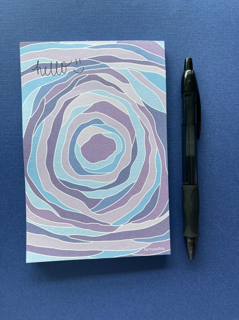 May include: A blue and purple patterned notepad with the text "hello :)" written on it. The notepad is on a blue surface with a black pen next to it. The notepad has the text "Fortuna Pins" at the bottom.