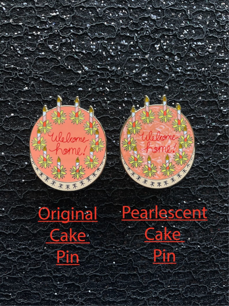 May include: Two round enamel pins shaped like cakes with the text Welcome home! in red script. The left pin has a solid peach surface, while the right pin features a pearlescent finish. Both pins have yellow candles and a decorative border.