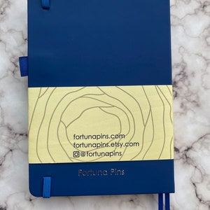 Op de afbeelding: Een donkerblauw notitieboek met een gele en witte band met een patroon. De band heeft de tekst "fortunapins.com", "fortunapins.etsy.com" en "@fortunapins". Het notitieboek heeft ook de tekst "Fortuna Pins" in gouden letters.