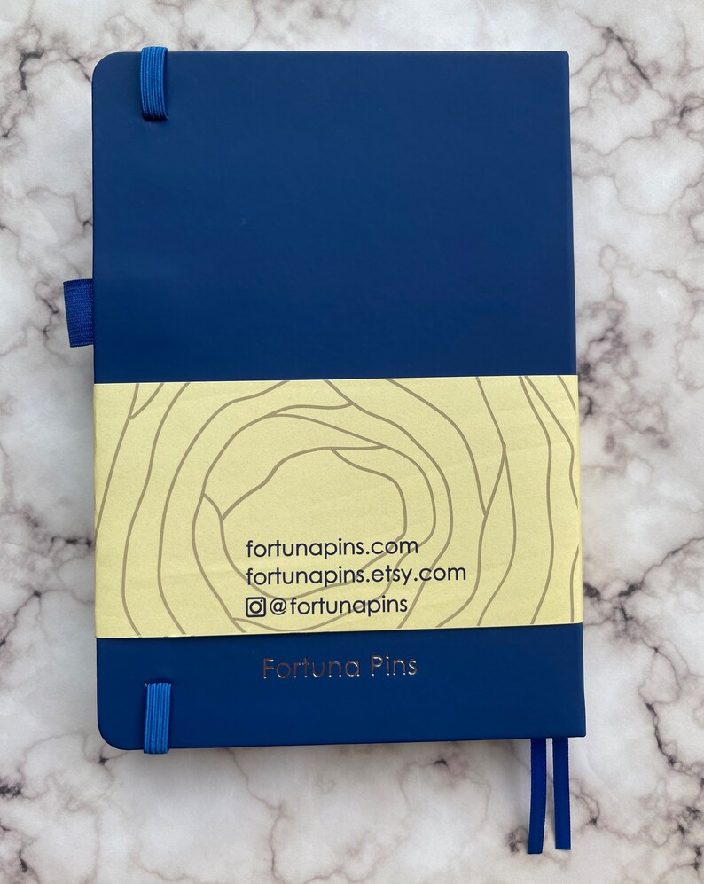 Op de afbeelding: Een donkerblauw notitieboek met een gele en witte band met een patroon. De band heeft de tekst "fortunapins.com", "fortunapins.etsy.com" en "@fortunapins". Het notitieboek heeft ook de tekst "Fortuna Pins" in gouden letters.
