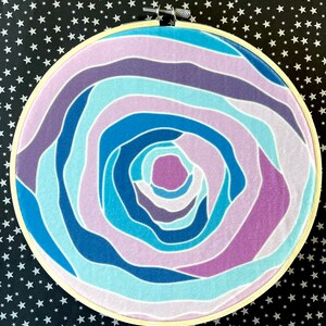 Puede incluir: Un aro de bordado circular con un dise&ntilde;o abstracto colorido. El dise&ntilde;o presenta l&iacute;neas en espiral en tonos de azul, morado y rosa.