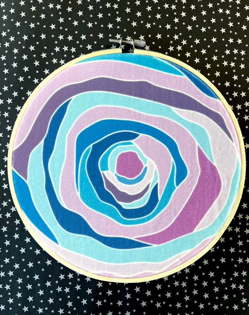 Puede incluir: Un aro de bordado circular con un dise&ntilde;o abstracto colorido. El dise&ntilde;o presenta l&iacute;neas en espiral en tonos de azul, morado y rosa.