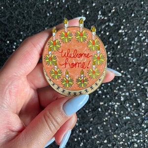 May include: A round enamel pin featuring a peach-colored swirled background with the text Welcome home! written in red script. The design is bordered by a ring of yellow flowers, each topped with a tall, thin candle with a gold glitter flame.