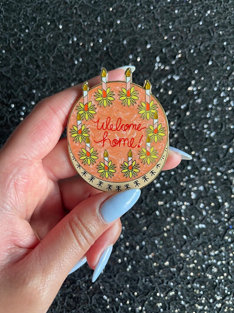 May include: A round enamel pin featuring a peach-colored swirled background with the text Welcome home! written in red script. The design is bordered by a ring of yellow flowers, each topped with a tall, thin candle with a gold glitter flame.