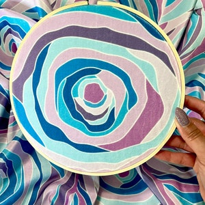 Puede incluir: Primer plano de un aro de bordado circular con un patr&oacute;n de tejido azul, morado y blanco en espiral. El tejido tiene una textura suave y aterciopelada.
