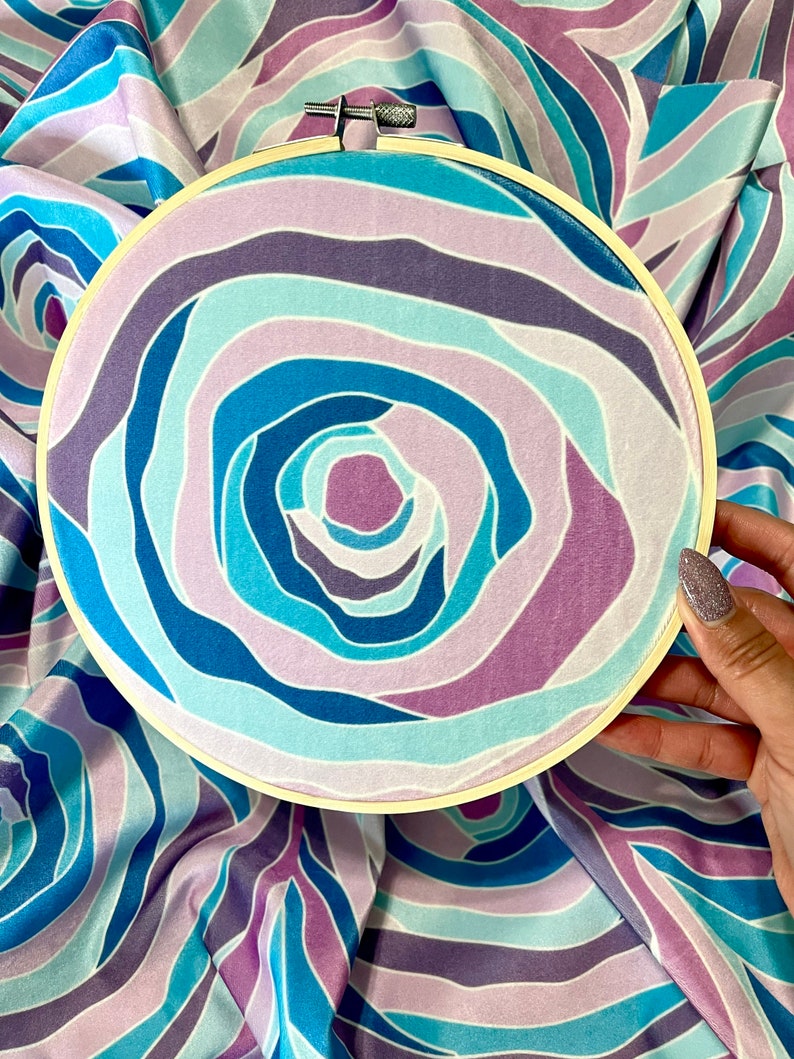 Puede incluir: Primer plano de un aro de bordado circular con un patr&oacute;n de tejido azul, morado y blanco en espiral. El tejido tiene una textura suave y aterciopelada.