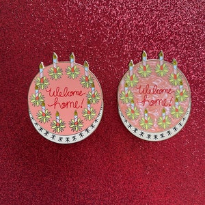 May include: Two round enamel pins shaped like cakes, each featuring the text "Welcome home!" in red script. The cakes are decorated with yellow flowers and candles. One pin has a solid pink background, while the other has a marbled pink finish.