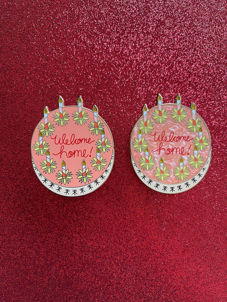 May include: Two round enamel pins shaped like cakes, each featuring the text "Welcome home!" in red script. The cakes are decorated with yellow flowers and candles. One pin has a solid pink background, while the other has a marbled pink finish.