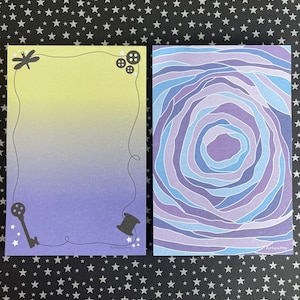 May include: Two notepads with different designs. The first notepad has a yellow to purple gradient background with a dragonfly, key, and spool of thread. The second notepad has a swirling blue and purple design. Both notepads have the text "Fortuna Pins" at the bottom.