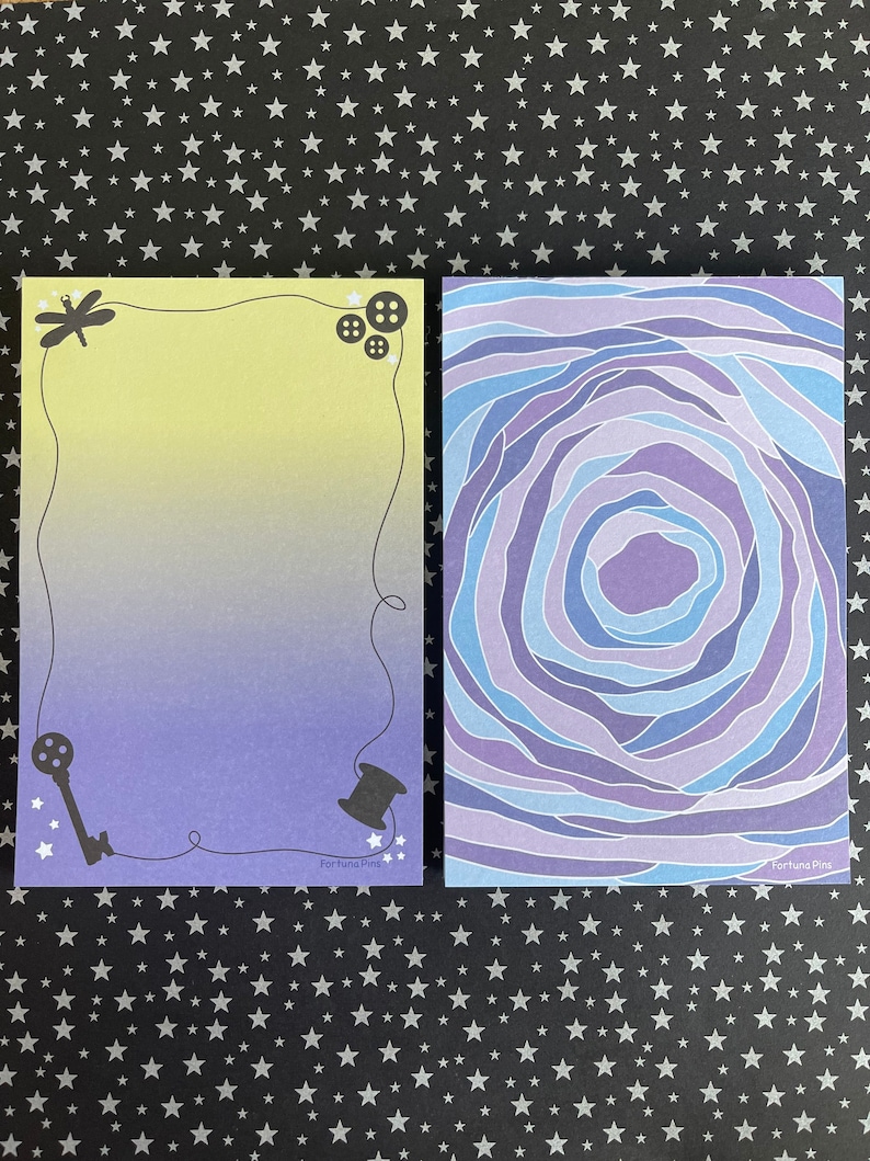 May include: Two notepads with different designs. The first notepad has a yellow to purple gradient background with a dragonfly, key, and spool of thread. The second notepad has a swirling blue and purple design. Both notepads have the text "Fortuna Pins" at the bottom.
