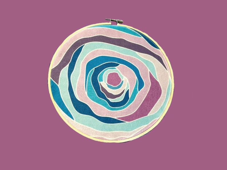 Puede incluir: Un aro de bordado circular con un patr&oacute;n de l&iacute;neas azules, rosas y blancas que se arremolinan. Las l&iacute;neas crean un efecto de espiral, que se asemeja a una flor o una galaxia.