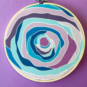 Puede incluir: Un aro de bordado circular con un patr&oacute;n de l&iacute;neas azules, moradas y blancas que se arremolinan. El patr&oacute;n est&aacute; centrado en el aro y crea un efecto de espiral.