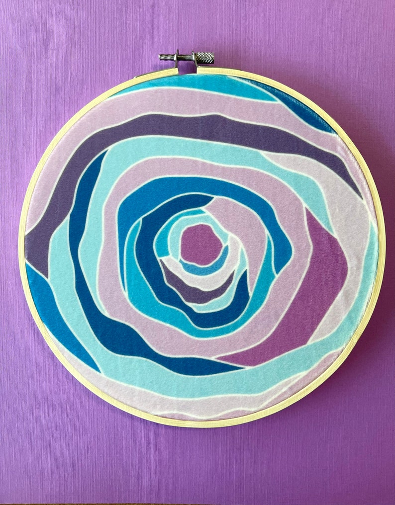 Puede incluir: Un aro de bordado circular con un patr&oacute;n de l&iacute;neas azules, moradas y blancas que se arremolinan. El patr&oacute;n est&aacute; centrado en el aro y crea un efecto de espiral.