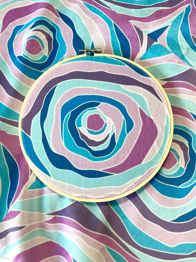 Puede incluir: Un aro de bordado circular con un inserto de tela que presenta un patr&oacute;n de rayas azules, moradas y rosas en espiral. La tela est&aacute; estirada sobre el aro.