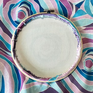 Puede incluir: Un aro de bordado de madera con una tela blanca estirada en el centro. El aro est&aacute; rodeado de una tela estampada en morado y azul.