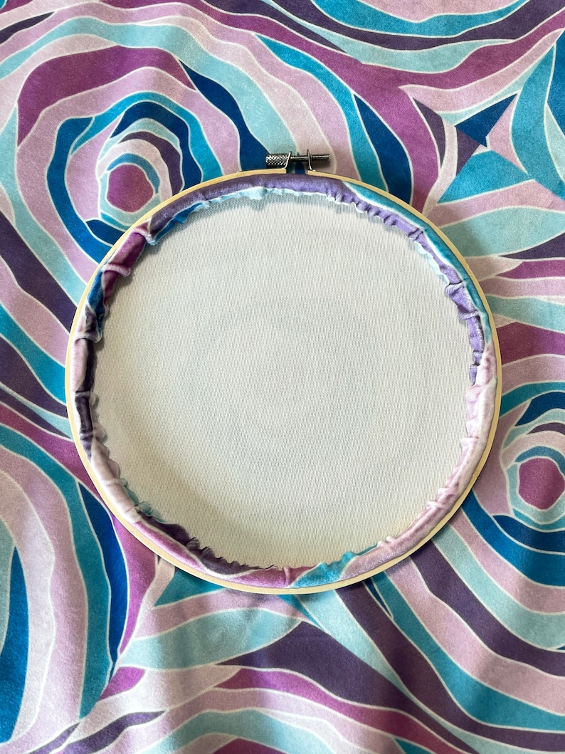 Puede incluir: Un aro de bordado de madera con una tela blanca estirada en el centro. El aro est&aacute; rodeado de una tela estampada en morado y azul.