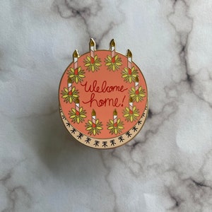 May include: A round enamel pin featuring a salmon-pink cake decorated with eight striped candles, each set in a yellow flower base. The text "Welcome home!" is written in red cursive in the center. The bottom edge has a black decorative border.