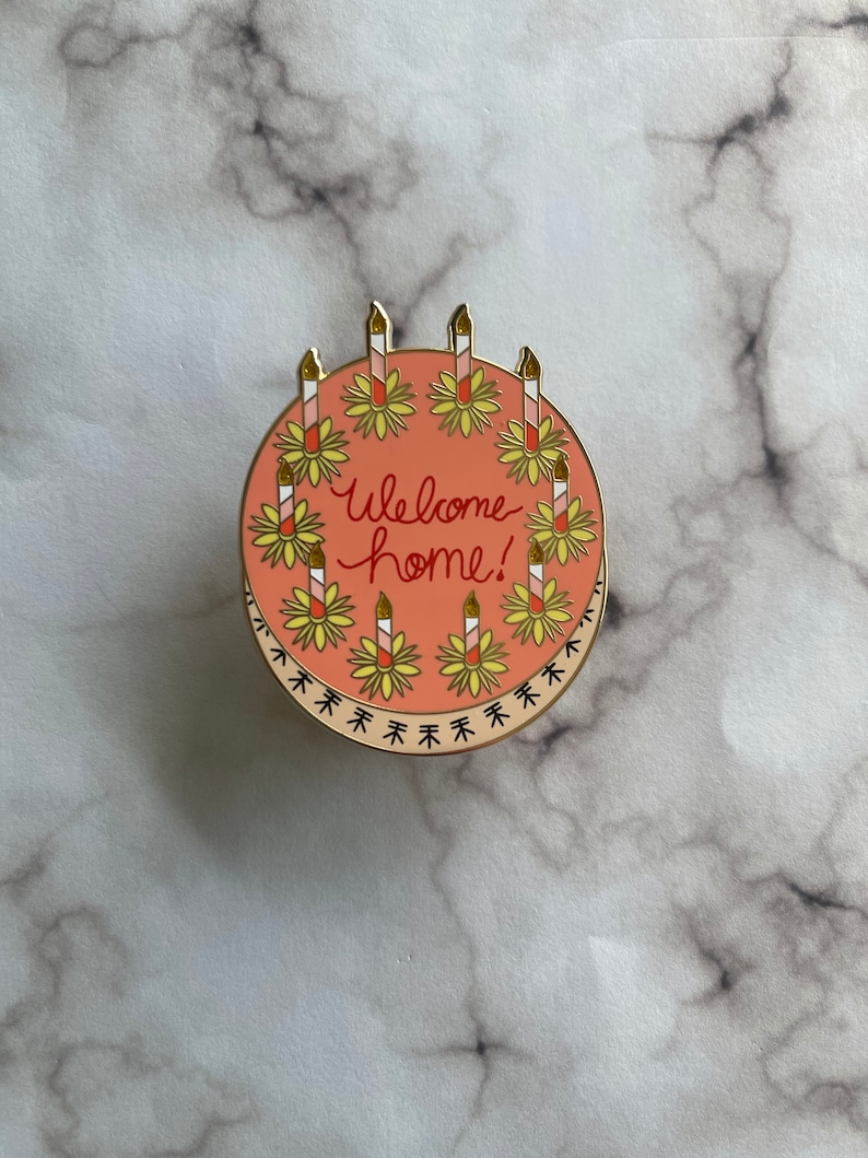 May include: A round enamel pin featuring a salmon-pink cake decorated with eight striped candles, each set in a yellow flower base. The text "Welcome home!" is written in red cursive in the center. The bottom edge has a black decorative border.