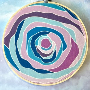 Puede incluir: Un aro de bordado circular con un patr&oacute;n de espiral en colores azul, morado y rosa. El patr&oacute;n recuerda a una rosa o una galaxia espiral.