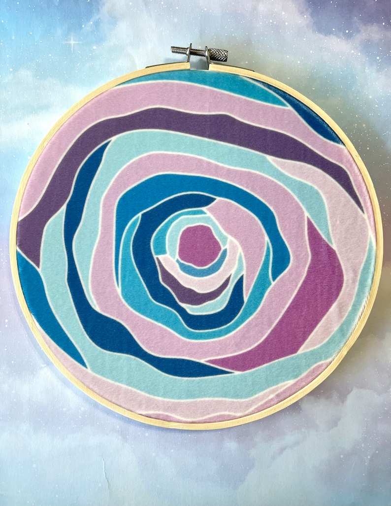Puede incluir: Un aro de bordado circular con un patr&oacute;n de espiral en colores azul, morado y rosa. El patr&oacute;n recuerda a una rosa o una galaxia espiral.