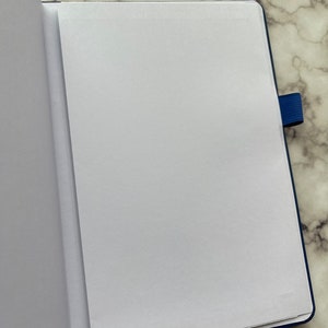 Op de afbeelding: Een open, blanco, blauw notitieboek met een witte pagina die naar voren is gekeerd. Het notitieboek heeft een blauwe elastieken sluiting.