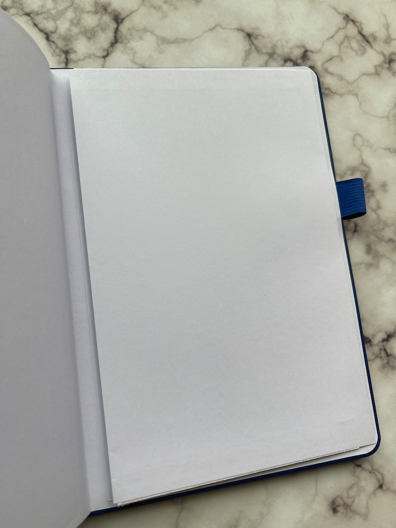 Op de afbeelding: Een open, blanco, blauw notitieboek met een witte pagina die naar voren is gekeerd. Het notitieboek heeft een blauwe elastieken sluiting.