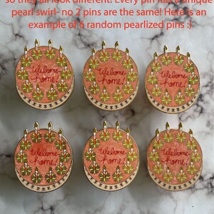 May include: Six round enamel pins shaped like cakes with a pearlescent pink swirled finish. Each pin features the text Welcome home! in red script, surrounded by yellow and white candles with gold metal outlines and a decorative border.