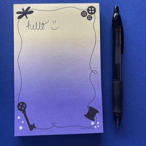 May include: A light blue and yellow notepad with a black border and various black illustrations including a dragonfly, buttons, a key, a spool of thread, and stars. The notepad has the text "hello" and a smiley face written on it. The text "Fortuna Pins" is printed at the bottom of the notepad.