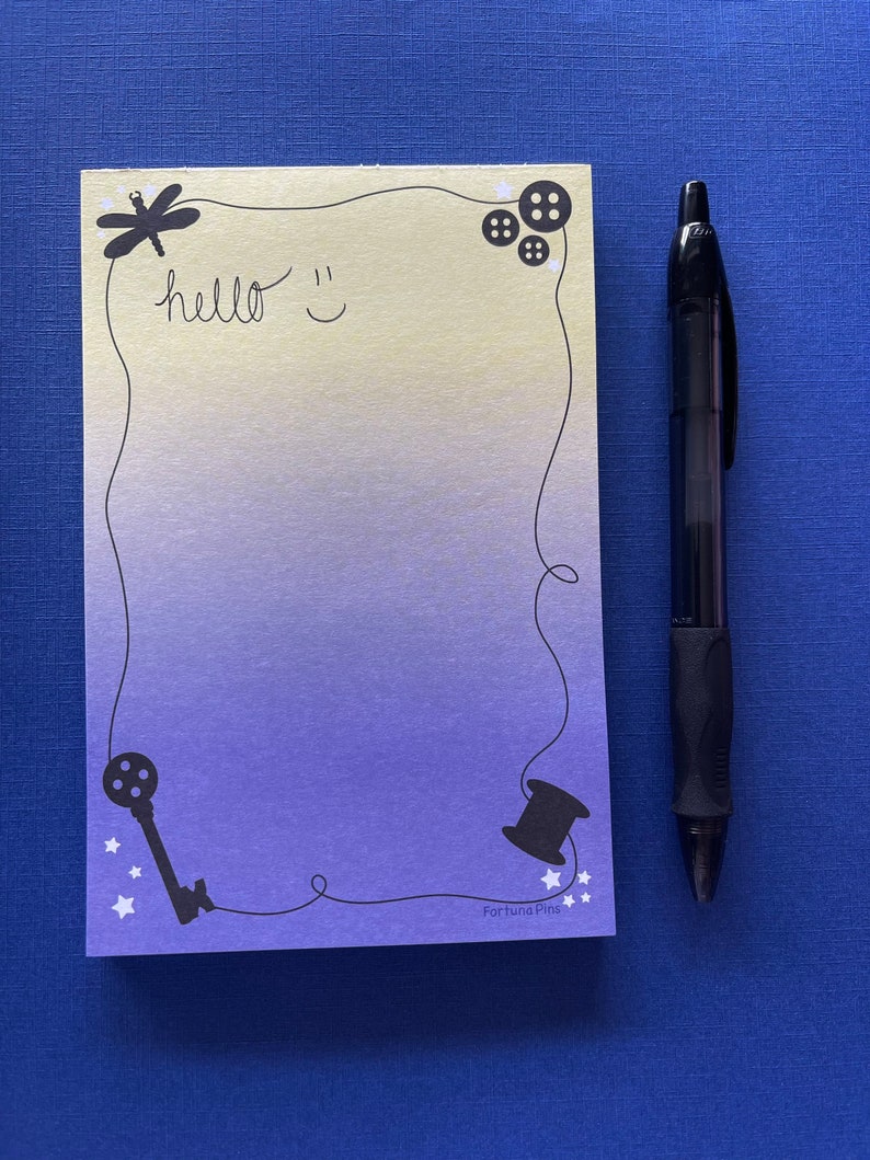 May include: A light blue and yellow notepad with a black border and various black illustrations including a dragonfly, buttons, a key, a spool of thread, and stars. The notepad has the text "hello" and a smiley face written on it. The text "Fortuna Pins" is printed at the bottom of the notepad.