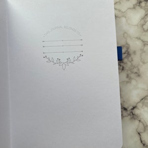 Op de afbeelding: Een wit dagboek met een grijze inscriptie die luidt "Dit dagboek behoort toe aan." De inscriptie is omgeven door een decoratieve rand van bladeren en wijnstokken.