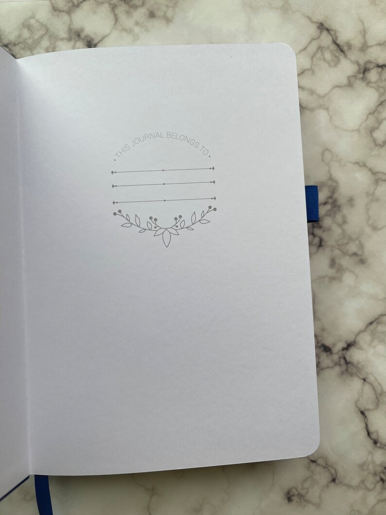 Op de afbeelding: Een wit dagboek met een grijze inscriptie die luidt "Dit dagboek behoort toe aan." De inscriptie is omgeven door een decoratieve rand van bladeren en wijnstokken.