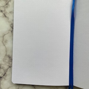 Op de afbeelding: Een wit notitieboek met een blauw lint als bladwijzer. Het notitieboek heeft een gestippeld raster op de pagina's.