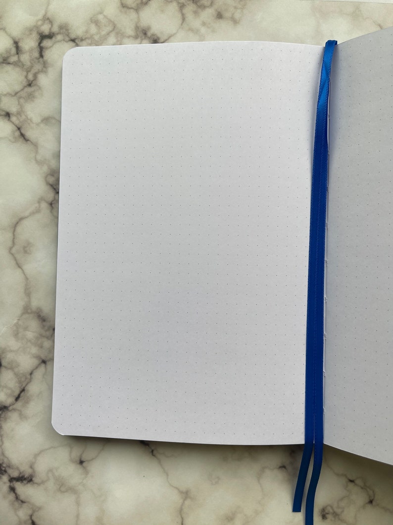 Op de afbeelding: Een wit notitieboek met een blauw lint als bladwijzer. Het notitieboek heeft een gestippeld raster op de pagina's.