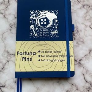 May include: A blue A5 bullet journal with a silver design on the front cover. The design features a key, a button, and a dragonfly. The text on the cover reads "Be Wise. Be Brave. Be Tricky." The journal is made of 160 GSM ultra thick paper and has 160 dot-grid pages.