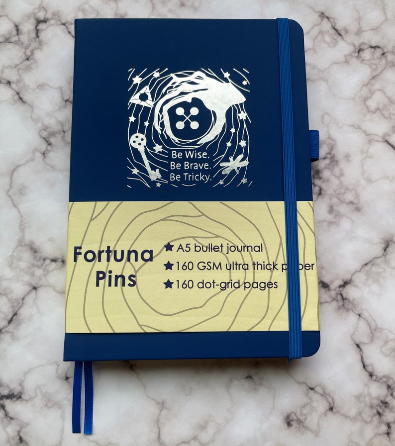 Op de afbeelding: Een blauwe A5 bullet journal met een zilveren ontwerp op de voorkant. Het ontwerp bevat een sleutel, een knoop en een libel. De tekst op de omslag luidt "Be Wise. Be Brave. Be Tricky." De journal is gemaakt van 160 GSM ultra dik papier en heeft 160 stippenrasterpagina's.