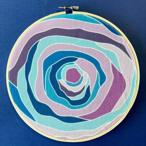 Puede incluir: Un aro de bordado circular con un dise&ntilde;o abstracto colorido. El dise&ntilde;o presenta l&iacute;neas en espiral en tonos de azul, morado y rosa.