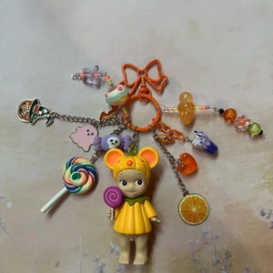 May include: A Halloween-themed keychain featuring a small figure in a yellow costume with mouse ears, holding a lollipop. The keychain includes charms of a ghost, candy, a slice of orange, and a bow. The keychain is attached to an orange ring.