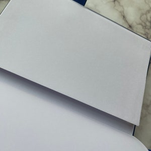 Op de afbeelding: Een blauw notitieboek met een witte omslag en een witte pagina die open is geslagen. Het notitieboek heeft een blauw lint als bladwijzer.
