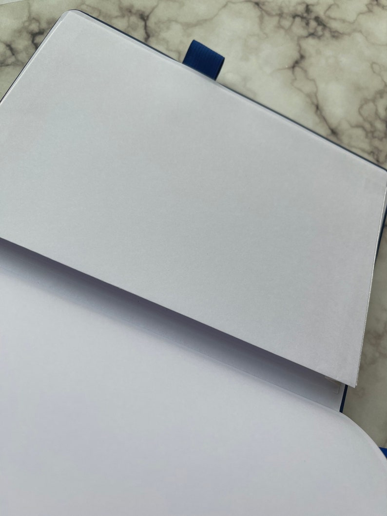 Op de afbeelding: Een blauw notitieboek met een witte omslag en een witte pagina die open is geslagen. Het notitieboek heeft een blauw lint als bladwijzer.