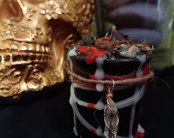 May include: A black candle with white and red wax drips, adorned with dried herbs and a clear crystal pendant. The candle is wrapped with twine and set against a gold skull background, creating a mystical aesthetic.