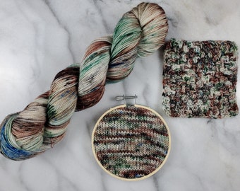 May include: A skein of yarn in shades of brown, green, and blue, with a crocheted square and a knitted swatch in the same colour scheme.