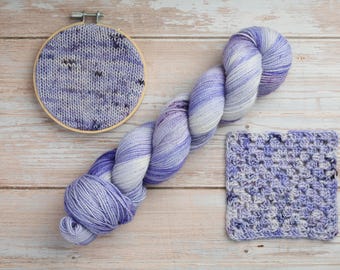 Könnte beinhalten: Ein Strang aus violett-weiß meliertem Garn, ein runder Stickrahmen aus Holz mit einem gestrickten violett-weißen Musterstück und ein quadratisches, gehäkeltes violett-weißes Stück mit strukturiertem Lochmuster.