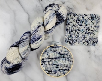 Könnte beinhalten: Eine blaue und weiße Garnrolle, ein gehäkeltes Quadrat und ein kleiner runder Stickrahmen mit einem gestrickten Muster darin.