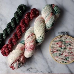 May include: An assortment of colorful yarn skeins in green, red, and cream, alongside a small embroidery hoop with a matching knitted design. The yarn is arranged on a marble surface, showcasing the textures and hues.