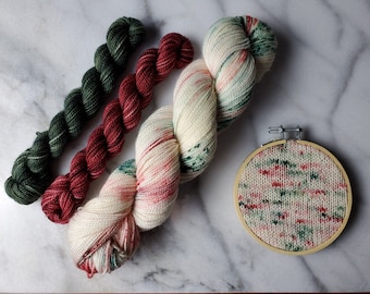 May include: An assortment of colorful yarn skeins in green, red, and cream, alongside a small embroidery hoop with a matching knitted design. The yarn is arranged on a marble surface, showcasing the textures and hues.