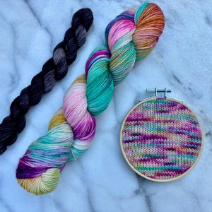 May include: An overhead shot features three items: a skein of black yarn, a colorful skein of yarn, and a small embroidery hoop with a knitted design. The colorful yarn has shades of pink, teal, and orange. The knitted design in the hoop has a mix of colors.