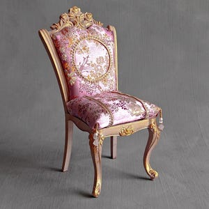 May include: A miniature chair featuring a gold-painted wooden frame with ornate carvings and cabriole legs. The seat and backrest are upholstered in a pink fabric with a metallic gold floral pattern, accented by hanging beaded tassels at the front.
